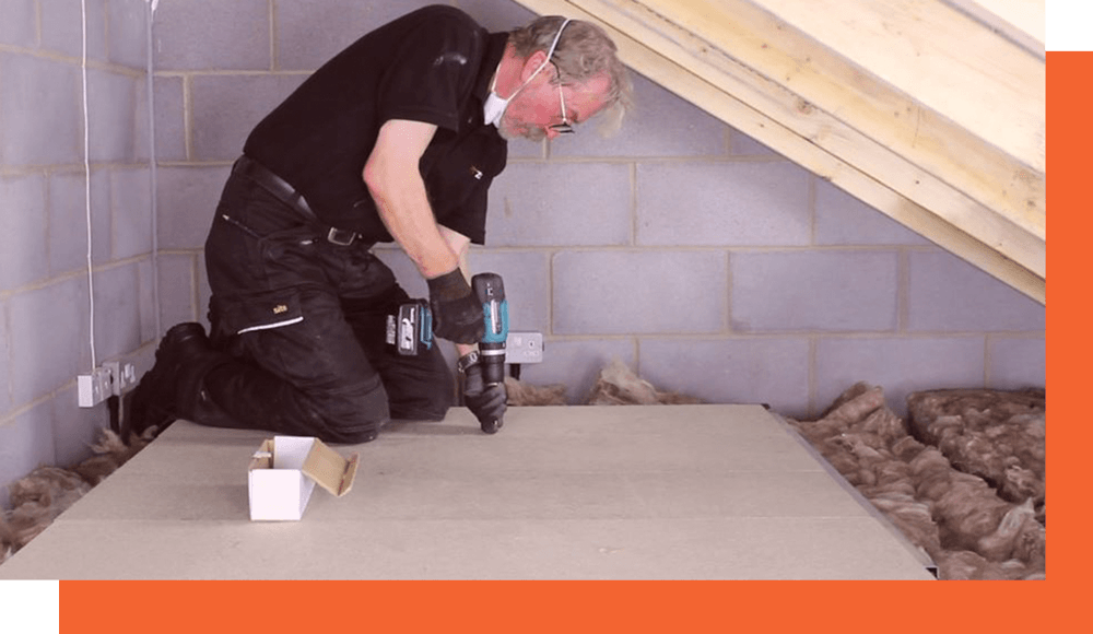 A man with a compact drill, drilling down attic floor boards.